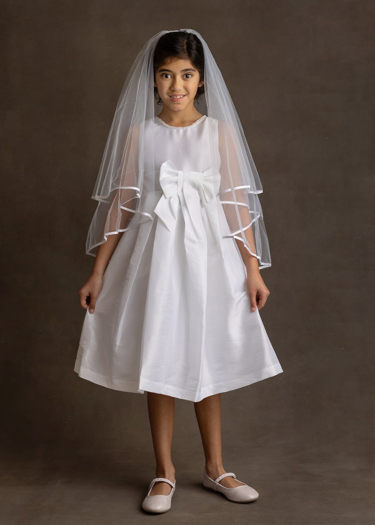 A young girl smiles softly at the camera in a Mabel and Honey Taffeta Bow Communion Dress with a beaded pearl neckline, a large waist bow, white veil, and light pink ballet flats against a plain brown background.