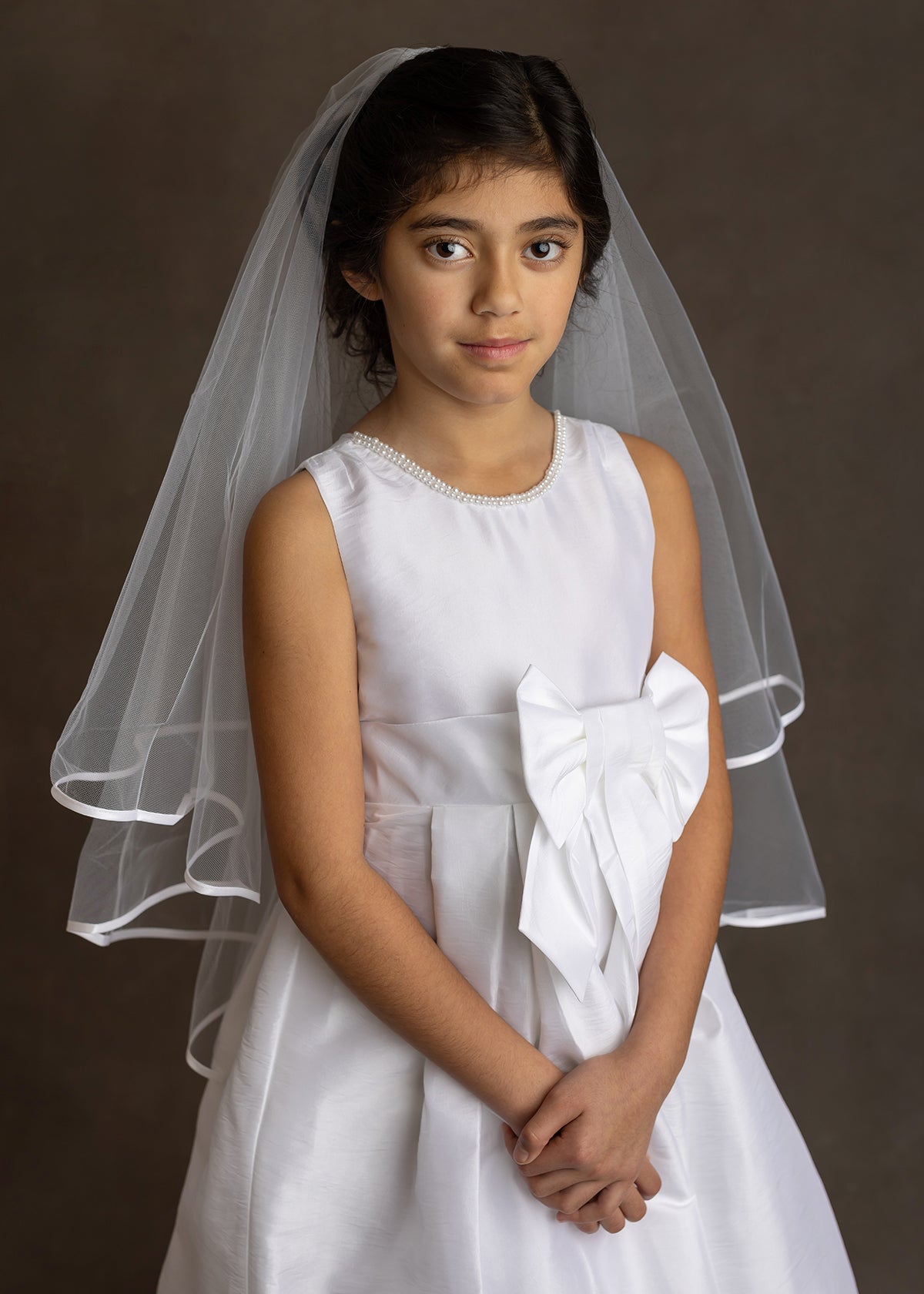 A young girl wears the Mabel and Honey Taffeta Bow Communion Dress with a beaded pearl neckline and sheer white veil, standing against a dark background with her hands folded.