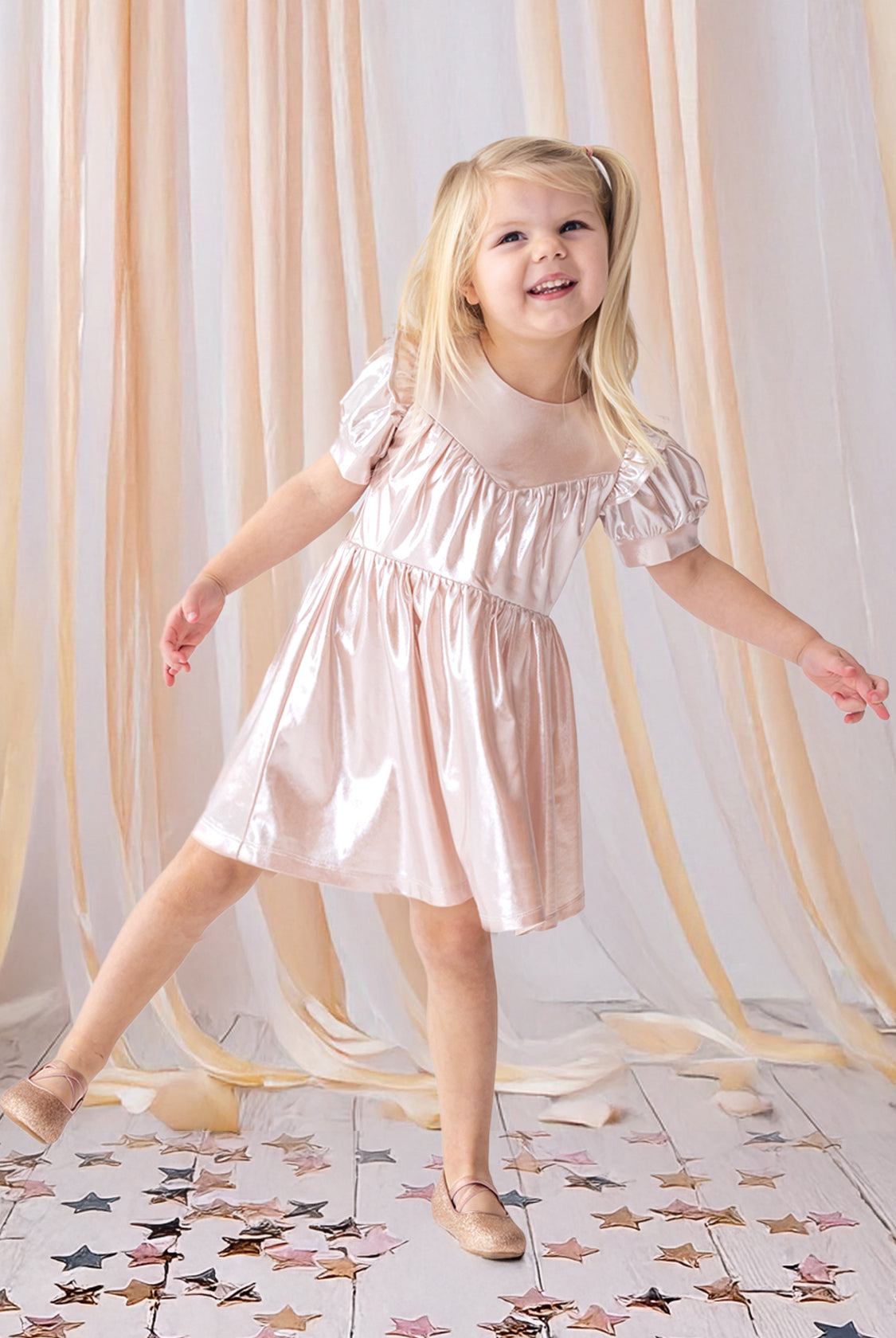 A young girl beams as she poses on one foot in the Isobella and Chloe Golden Glow Pink Dress, featuring shiny metallic pink fabric and puffed short sleeves, with matching shoes. She stands on a white floor amid gold and pink star confetti and peach drapes.