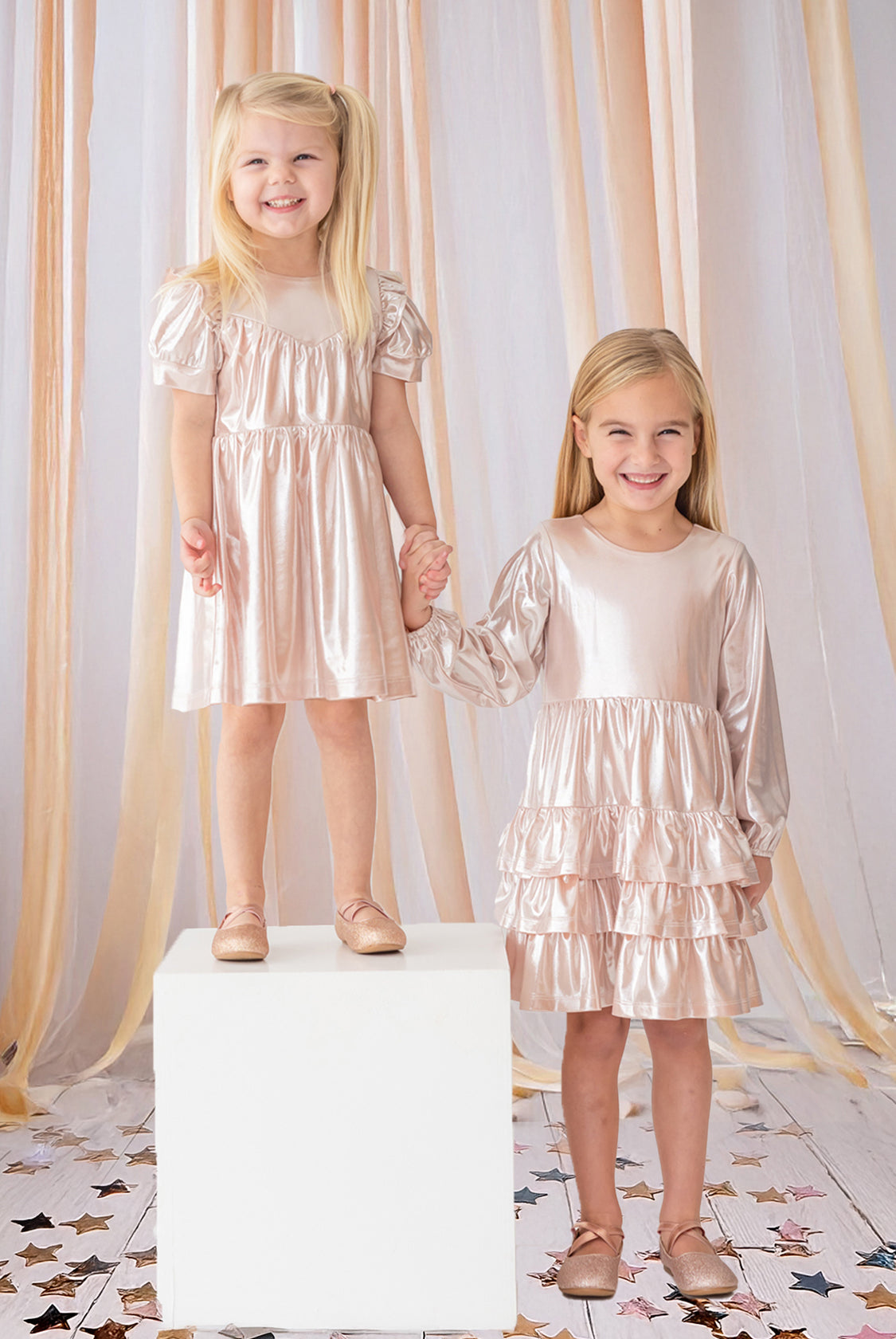 Two young girls hold hands and smile, both wearing Isobella and Chloe’s Golden Glow Pink Dresses with puffed sleeves and matching shoes. One girl stands on a white cube, surrounded by peach ribbons and star confetti for a magical touch.