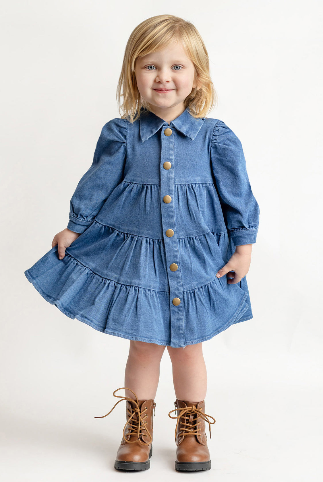 A young blonde-haired child wears the Mabel and Honey Denim Days Tiered Dress—a blue, long-sleeve, button-down denim dress—paired with brown lace-up boots, smiling while standing against a plain white background.