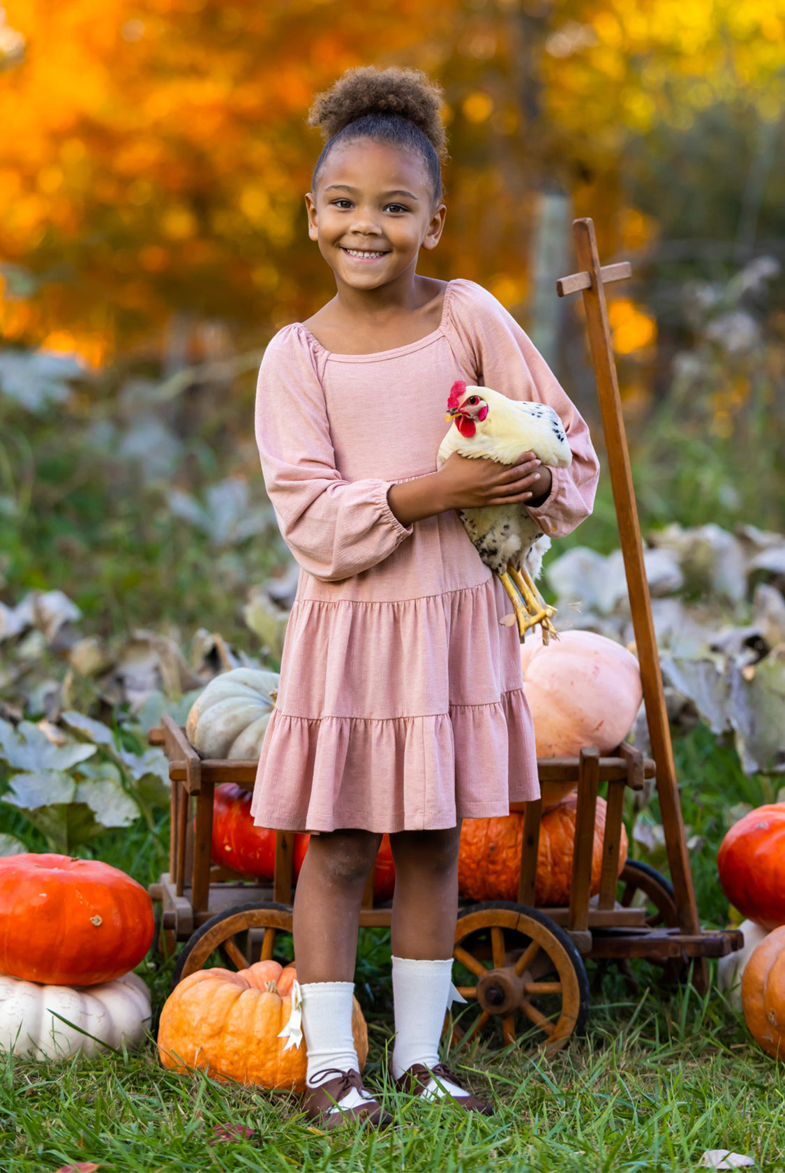 A smiling girl in the Mabel and Honey Flower Mae Pink Dress stands outdoors holding a chicken, surrounded by pumpkins and autumn foliage, with a wooden cart behind her for a cozy fall vibe.