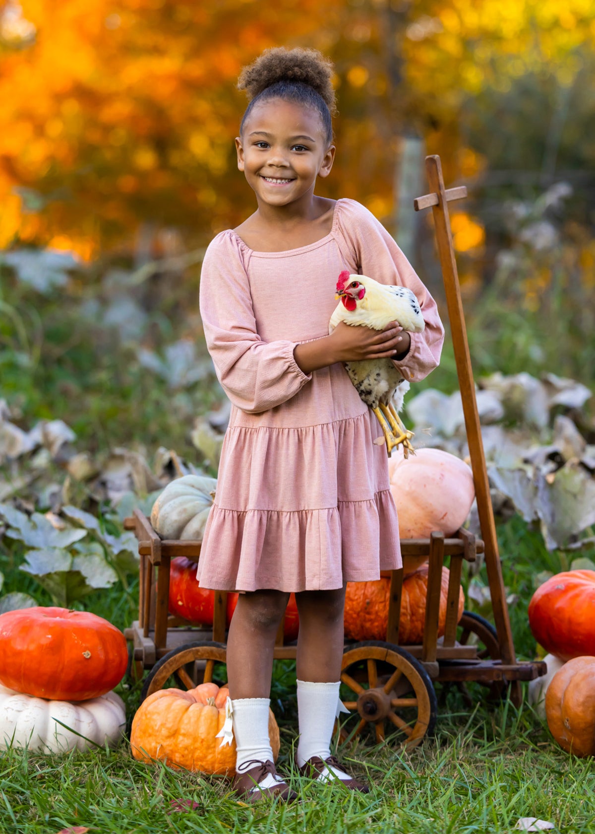 A smiling girl in the Mabel and Honey Flower Mae Pink Dress stands outdoors holding a chicken, surrounded by pumpkins and autumn foliage, with a wooden cart behind her for a cozy fall vibe.