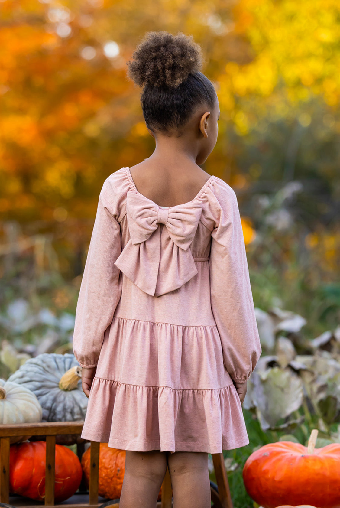 A young girl with curly hair in a bun stands outdoors among pumpkins and autumn leaves, wearing the Flower Mae Pink Dress by Mabel and Honey, featuring a large bow on the back.