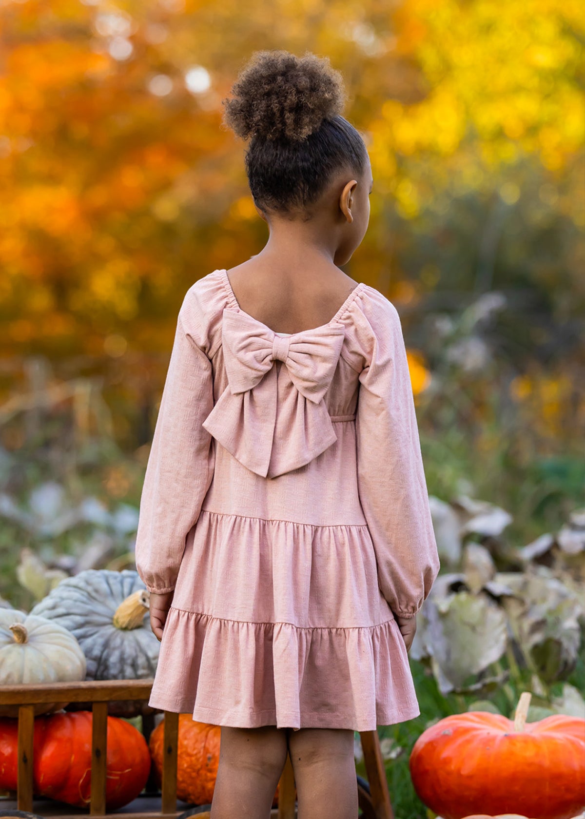 A young girl with curly hair in a bun stands outdoors among pumpkins and autumn leaves, wearing the Flower Mae Pink Dress by Mabel and Honey, featuring a large bow on the back.