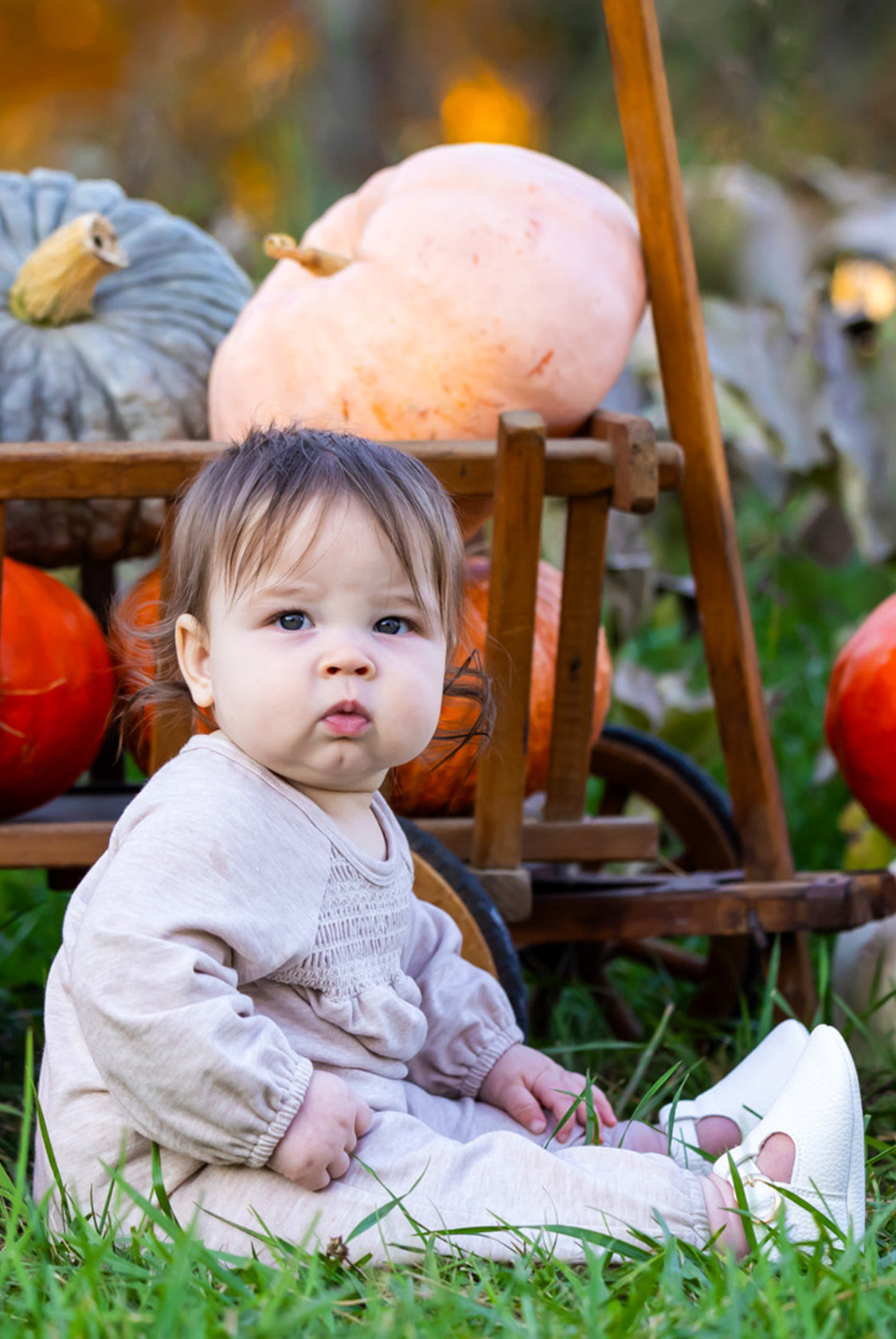 A baby wears the Millie Romper by Mabel and Honey while sitting on grass before a wooden cart of pumpkins and gourds, creating a cozy autumn scene with seasonal hues and greenery.