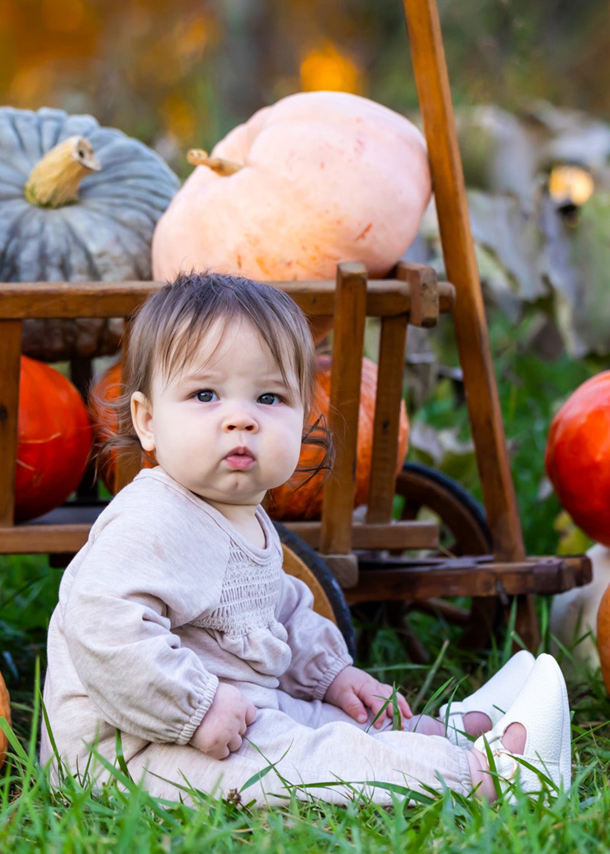 A baby wears the Millie Romper by Mabel and Honey while sitting on grass before a wooden cart of pumpkins and gourds, creating a cozy autumn scene with seasonal hues and greenery.