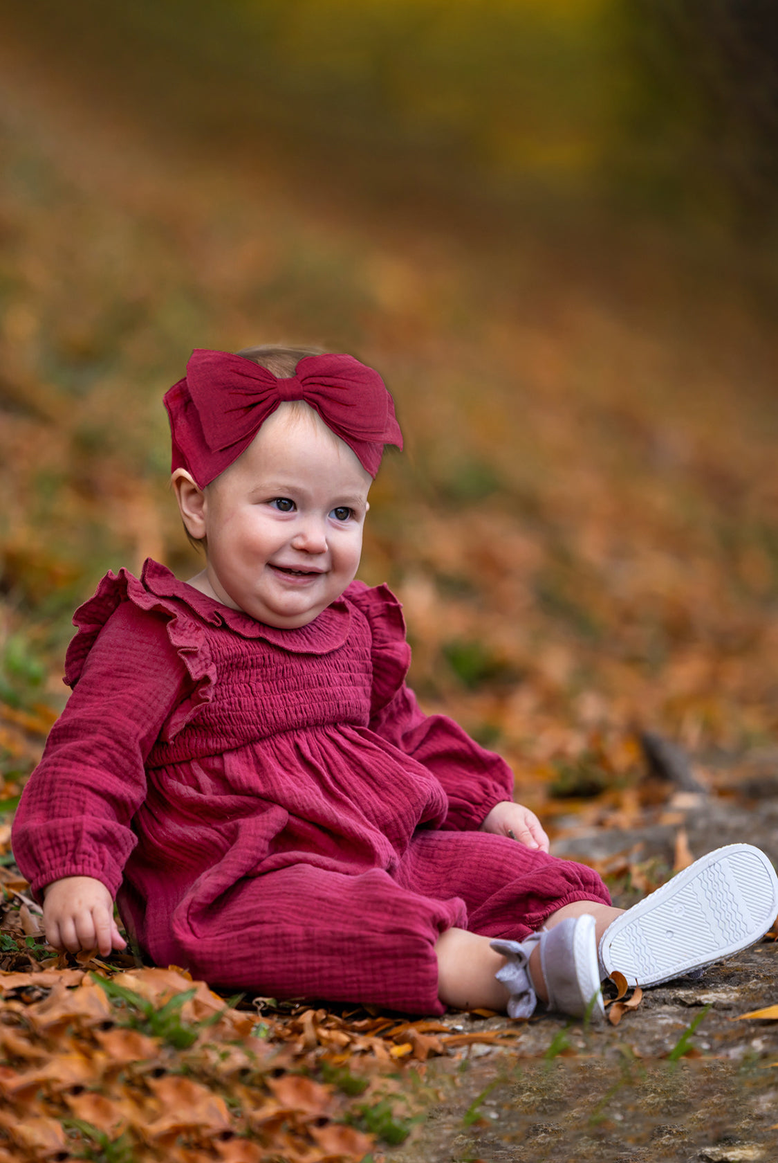 A smiling baby wears the Mabel and Honey Autumn Romper with a ruffled collar and matching headband, sitting on autumn leaves with trees softly blurred in the background.