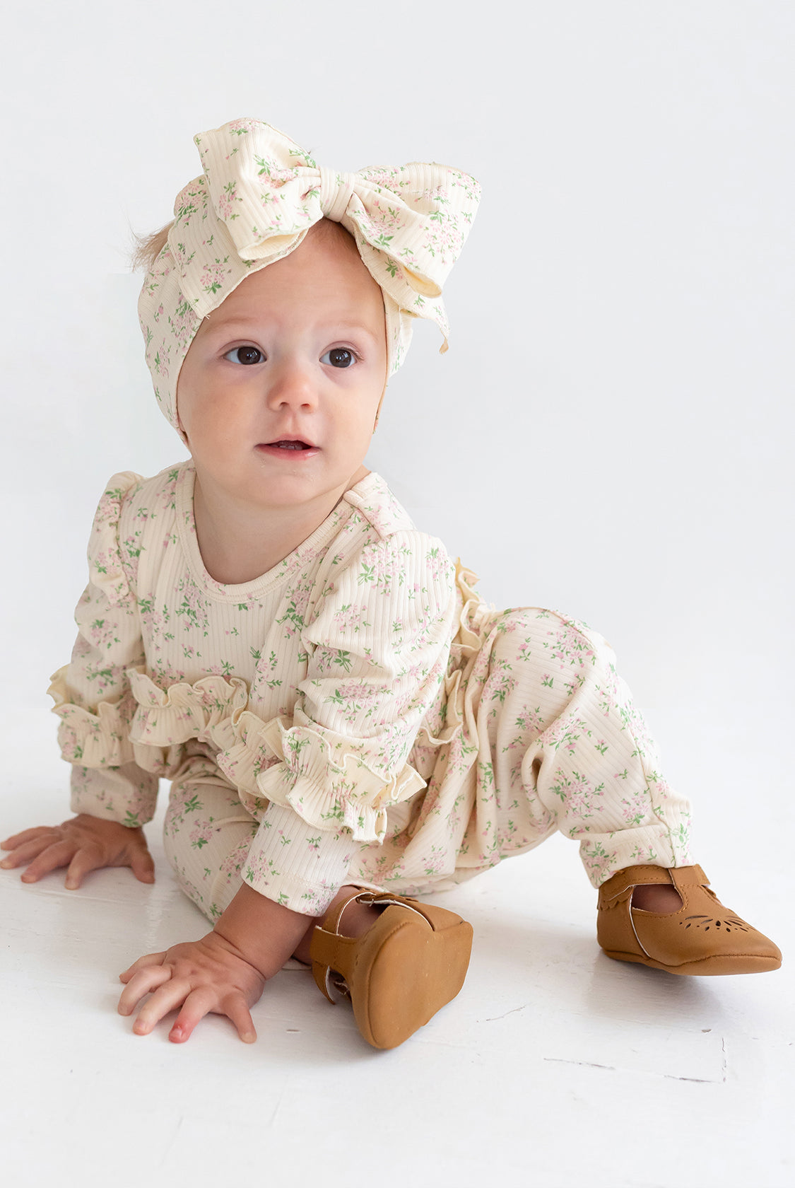 A baby wearing the Mabel and Honey "Breakfast in Bed" cream floral romper with ruffled sleeves, a matching large bow headband, and brown shoes sits on a white floor, gazing slightly to the side.