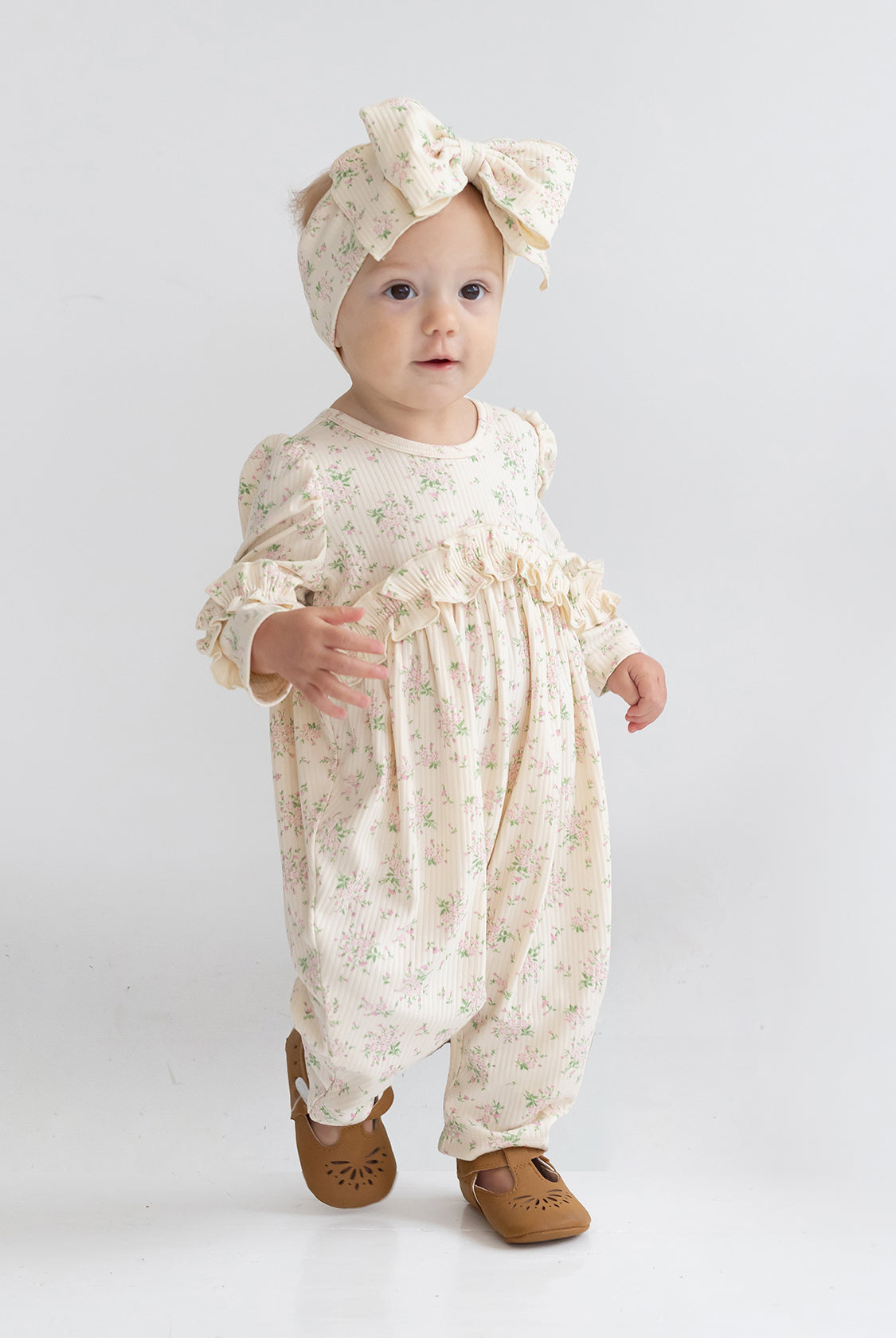 A toddler wears the Mabel and Honey "Breakfast in Bed Romper," a cream floral outfit with ruffled sleeves, paired with a matching large bow headband and brown shoes, standing against a plain white background.