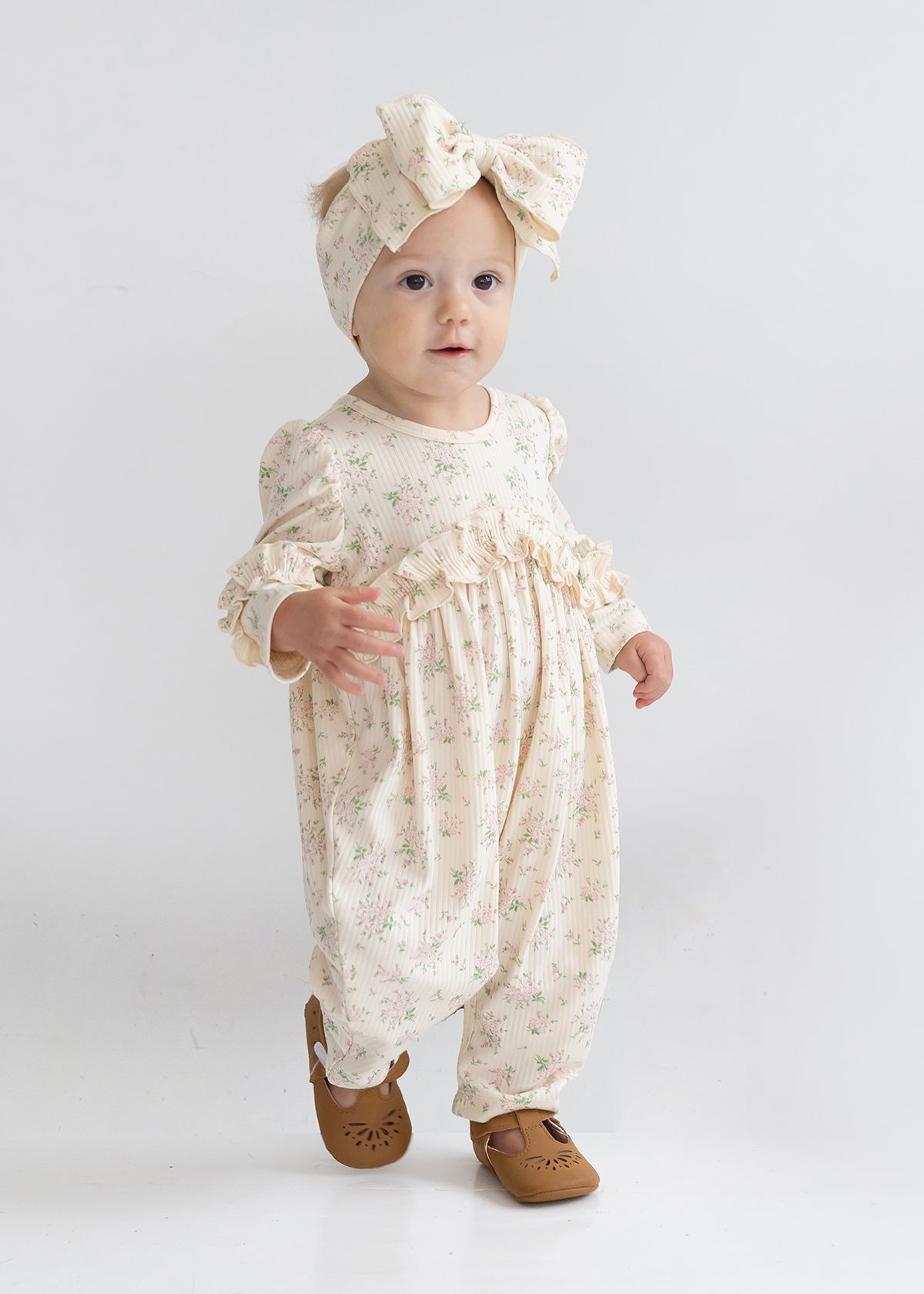 A toddler wears the Mabel and Honey "Breakfast in Bed Romper," a cream floral outfit with ruffled sleeves, paired with a matching large bow headband and brown shoes, standing against a plain white background.