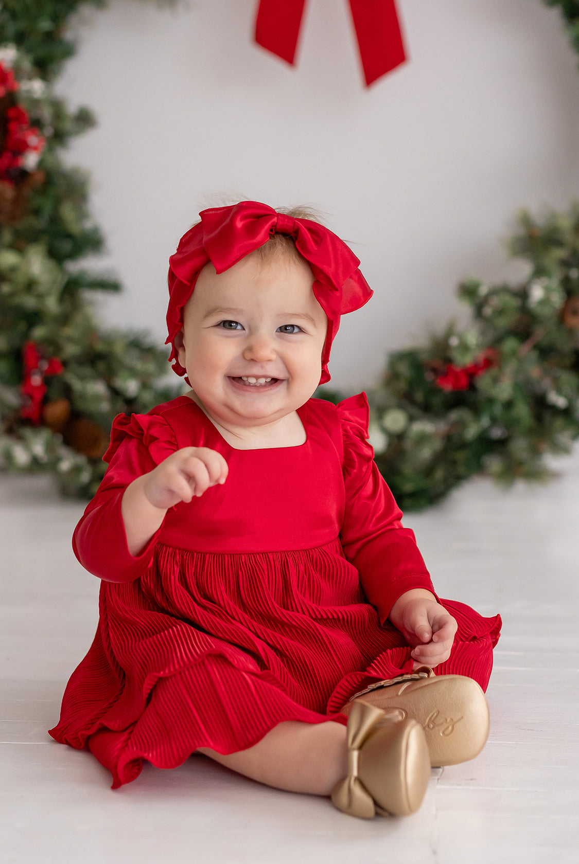 A smiling baby girl wearing the Isobella and Chloe Merry 'n Bright Red Velvet Dress and matching headband sits by a festive wreath. She pairs the outfit with gold shoes and reaches forward with one hand.