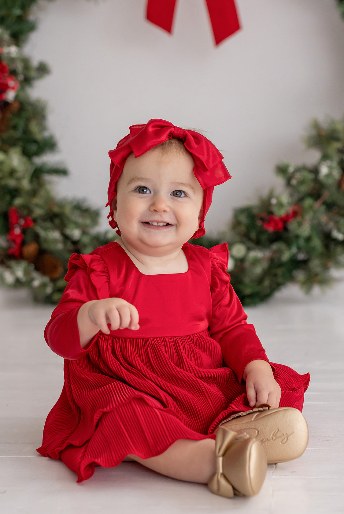 A smiling baby in the Merry 'n Bright Red Velvet Dress by Isobella and Chloe and a matching headband sits before a festive holiday wreath, wearing gold shoes labeled "Baby." Greenery on the white background creates a cheerful holiday scene.