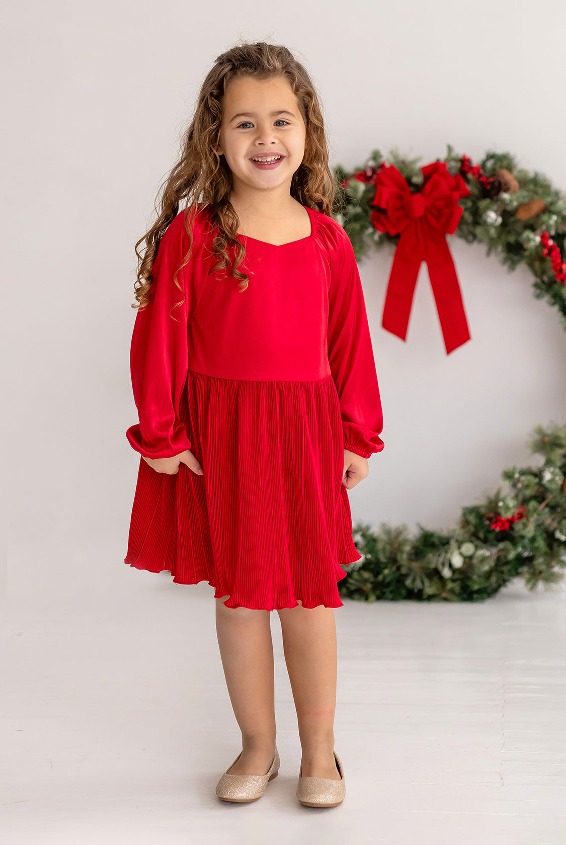 A young girl with long curly hair smiles indoors, wearing the Isobella and Chloe Merry n Bright Velvet Dress—red with long sleeves, natural waist, and pleated skirt—plus beige shoes, standing in front of a large wreath with a red bow.