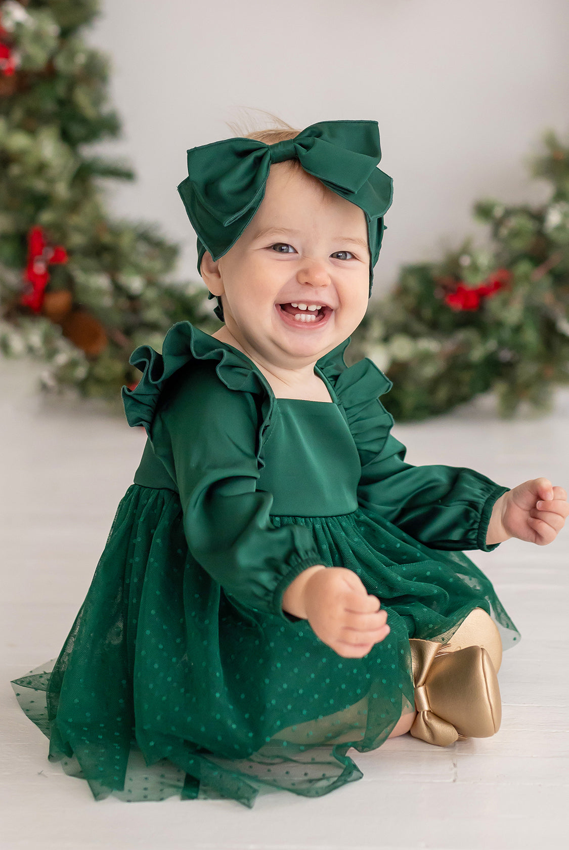 A smiling baby wears the Isobella and Chloe Mistletoe Emerald Dress with a matching headband and gold shoes, surrounded by holiday wreaths with red ribbons—perfect for festive celebrations.