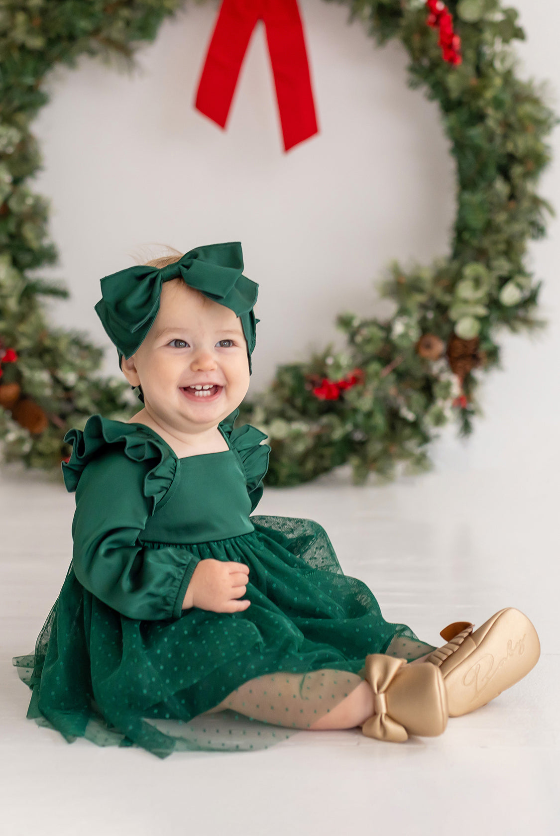A smiling baby girl wears the Isobella and Chloe Mistletoe Emerald Dress with a matching bow, gold shoes, and sheer polka-dot stockings as she sits before a festive wreath—perfect attire for holiday celebrations.