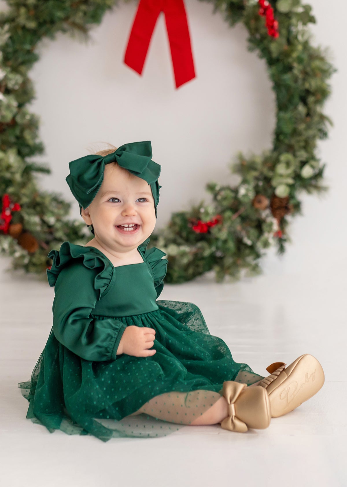 A smiling baby girl wears the Isobella and Chloe Mistletoe Emerald Dress with a matching bow, gold shoes, and sheer polka-dot stockings as she sits before a festive wreath—perfect attire for holiday celebrations.