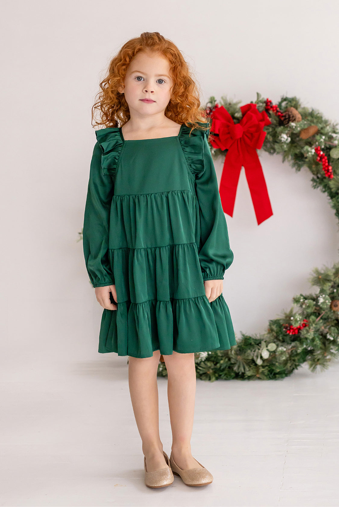 A young girl with curly red hair wears the Isobella and Chloe Mistletoe Emerald Green Tiered Dress and beige shoes, standing indoors on a white floor. A festive wreath with a red bow hangs in the background.