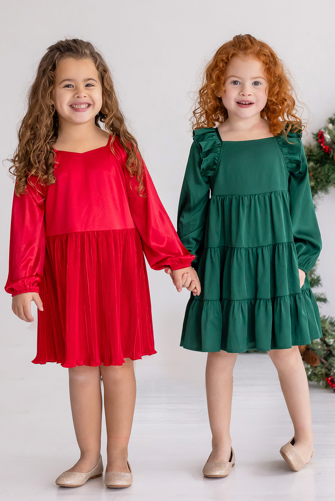 Two young girls stand side by side, smiling and holding hands. One wears a red holiday party dress, while the other wears the Isobella and Chloe Mistletoe Emerald Green Tiered Dress. Both have curly hair, beige shoes, and a festive wreath in the background.