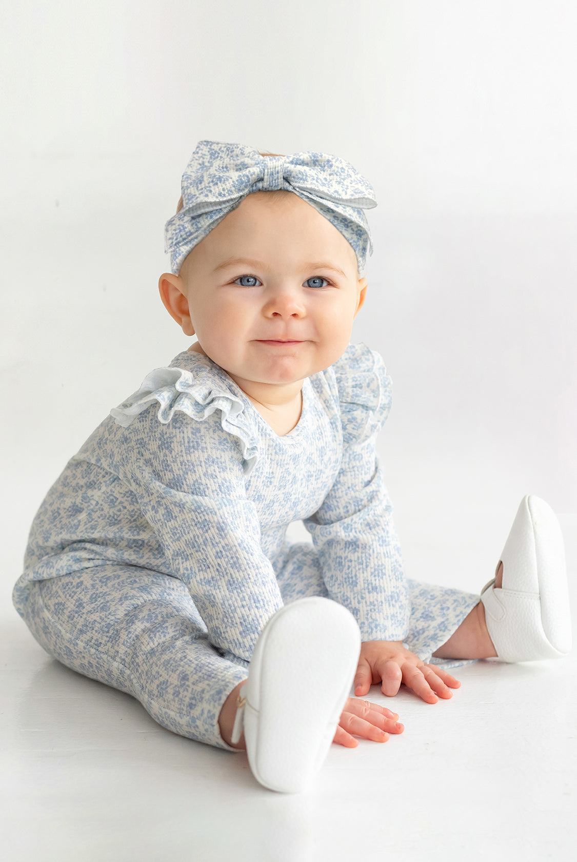 A baby wears the Mabel and Honey "Hannah Romper," featuring a blue and white floral print with ruffled shoulders, paired with a matching bow headband and white shoes while sitting on a white floor, smiling up.