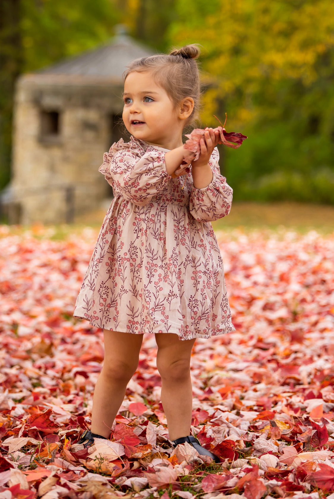 A young girl wears the Mabel and Honey Autumn Floral Dress Long Sleeve as she stands on fallen autumn leaves, smiling and looking to the side while holding red leaves. Blurred trees and a stone structure are in the background.
