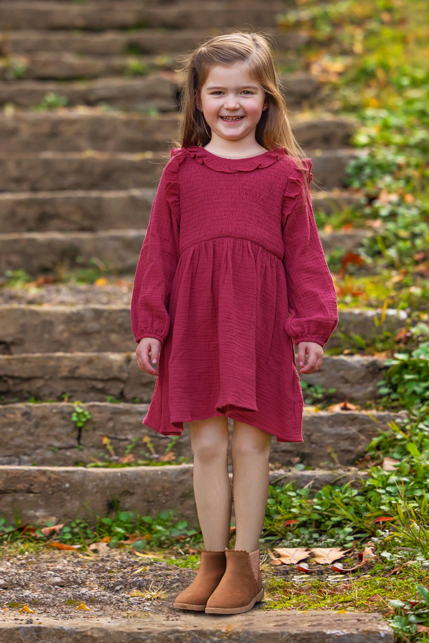 A young girl with long brown hair smiles brightly outdoors, wearing the Mabel and Honey Autumn Dress Red with ruffle details, surrounded by greenery and fallen leaves on stone steps.