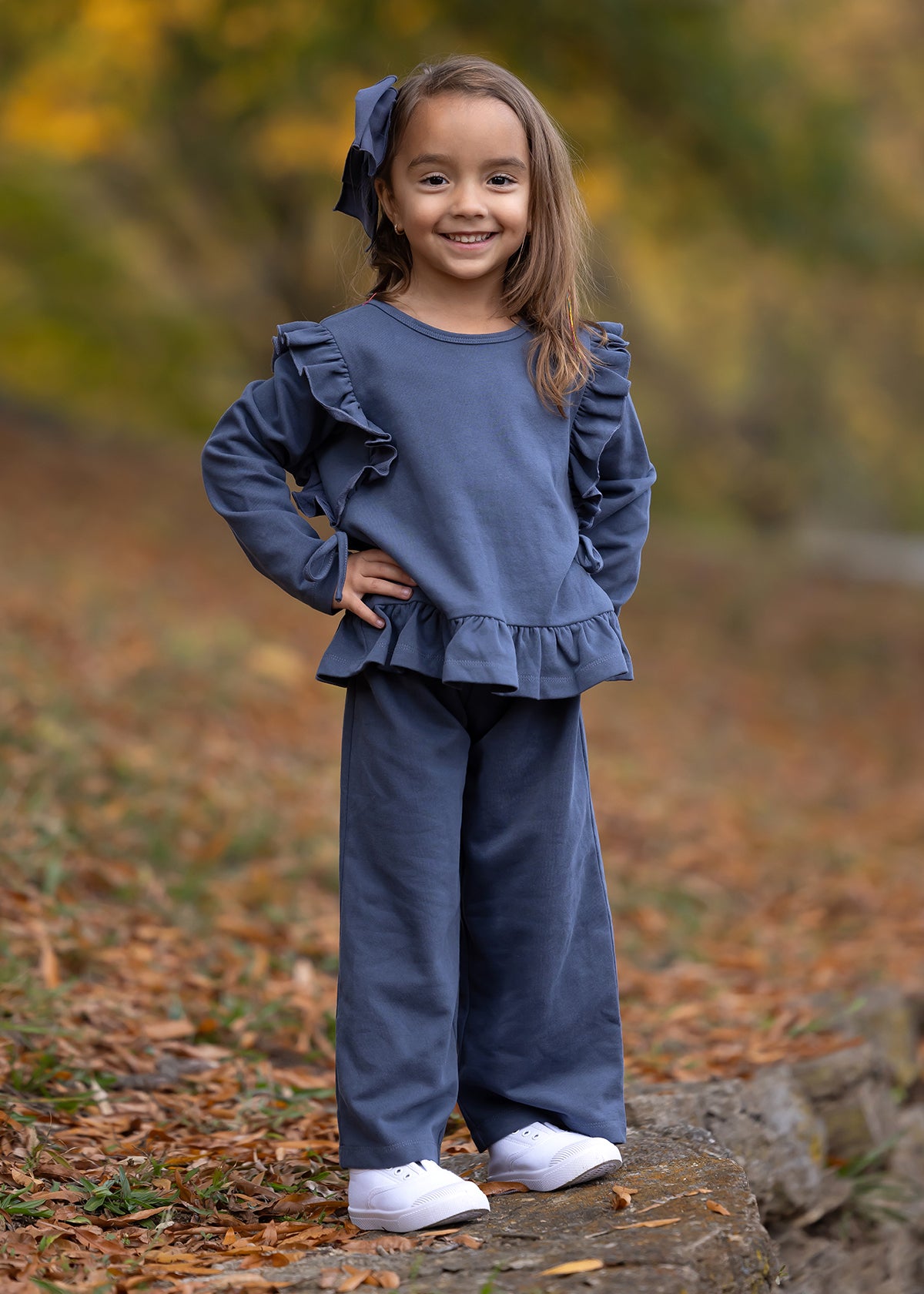 A young girl smiles outdoors on a leaf-strewn path, wearing the Mabel and Honey Robyn Two Piece Set with a ruffled peplum top, wide-leg pants, white shoes, and a big blue hair bow. Fall leaves blur in the background.