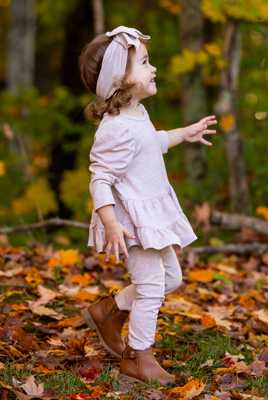 A young child wears the Millie Two Piece Set by Mabel and Honey, a light pink long sleeve outfit, paired with brown boots, walking through fallen autumn leaves with vibrant trees in the background.