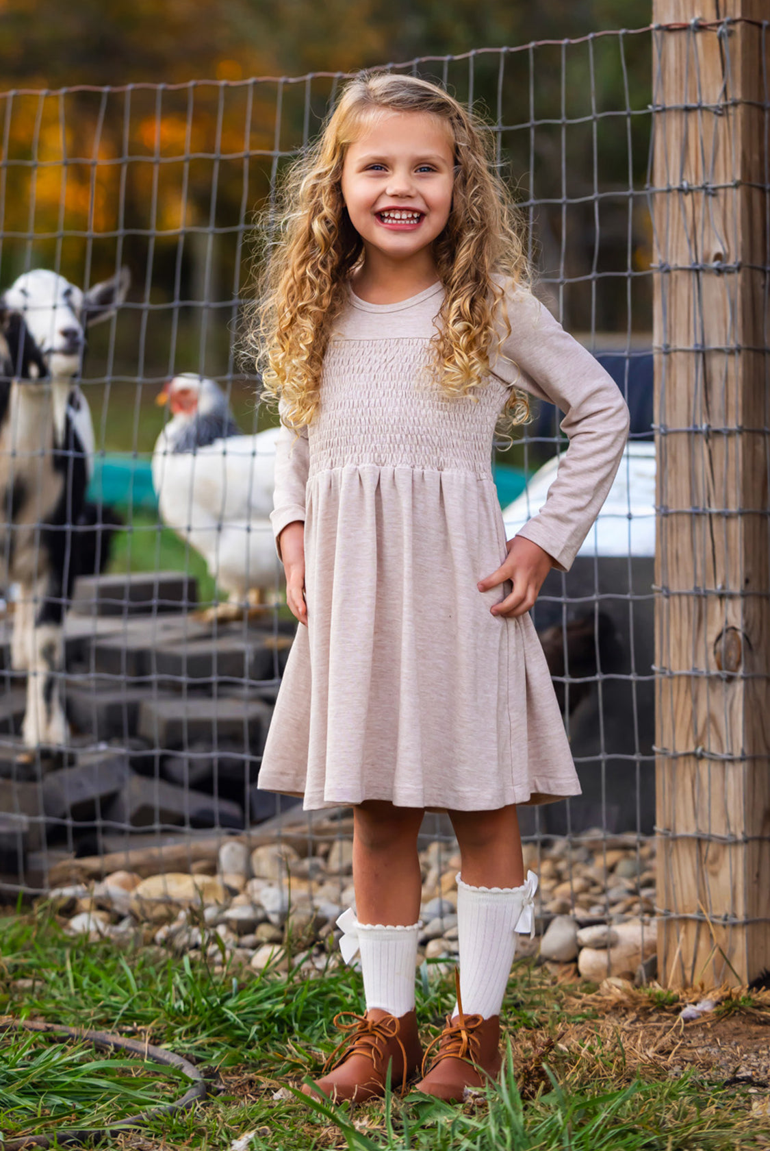 A young girl with curly blonde hair smiles in front of a wire fence with goats and a white chicken, wearing the Mabel and Honey Millie Natural Waist Dress, long socks, and brown shoes.