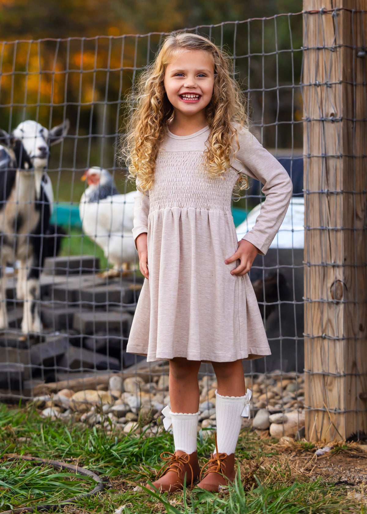 A young girl with curly blonde hair smiles in front of a wire fence with goats and a white chicken, wearing the Mabel and Honey Millie Natural Waist Dress, long socks, and brown shoes.