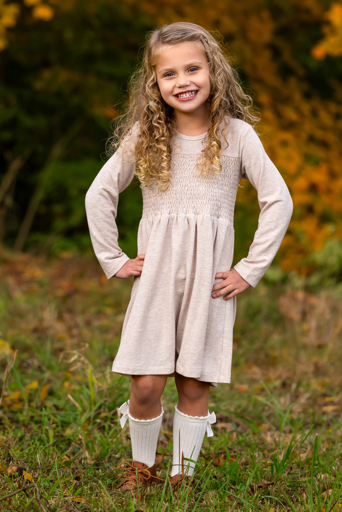 A smiling young girl stands on grass with her hands on her hips, wearing the Mabel and Honey Millie Natural Waist Dress, knee-high white socks with bows, and brown shoes. Autumn foliage is softly blurred in the background.