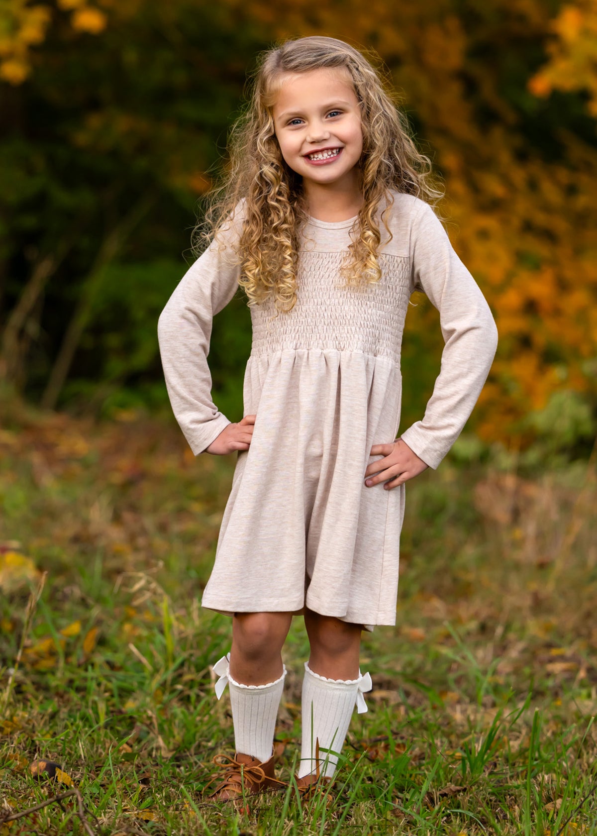 A smiling young girl stands on grass with her hands on her hips, wearing the Mabel and Honey Millie Natural Waist Dress, knee-high white socks with bows, and brown shoes. Autumn foliage is softly blurred in the background.