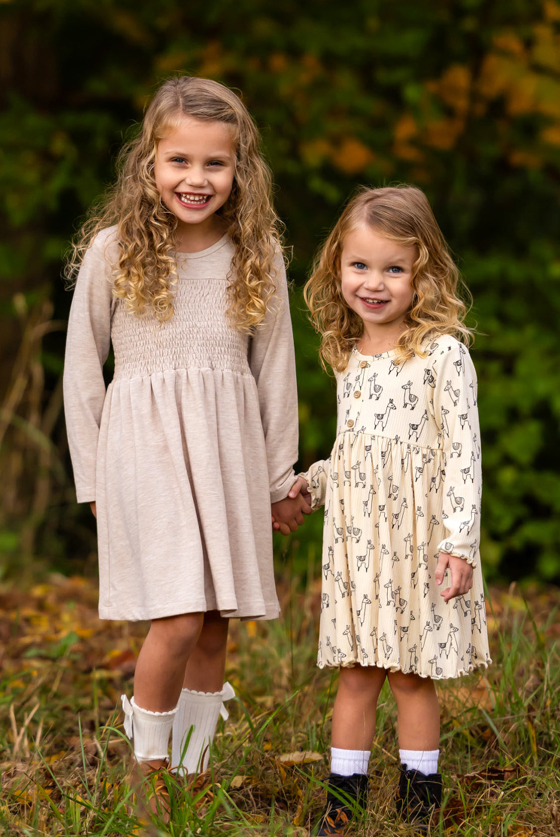 Two young girls with curly hair stand outdoors, holding hands and smiling. Both wear the Millie Natural Waist Dress from Mabel and Honey with ankle socks, surrounded by greenery and autumn foliage.