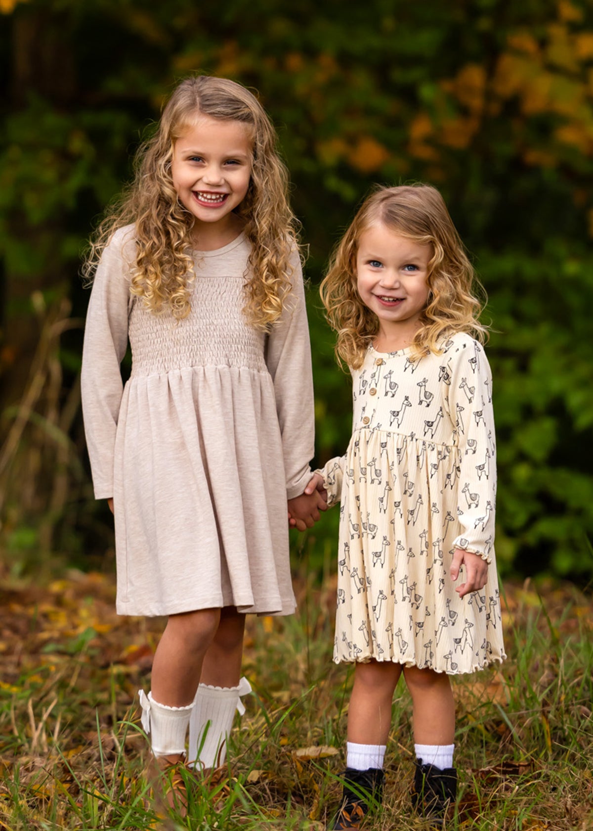 Two young girls with curly hair stand outdoors, holding hands and smiling. Both wear the Millie Natural Waist Dress from Mabel and Honey with ankle socks, surrounded by greenery and autumn foliage.