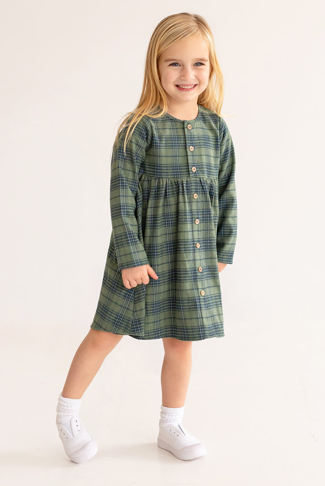 A young girl with long blonde hair smiles in the Cozy Creatures Dress by Mabel and Honey, featuring a green plaid pattern, long sleeves, and wooden buttons, paired with white socks and shoes on a plain light background.