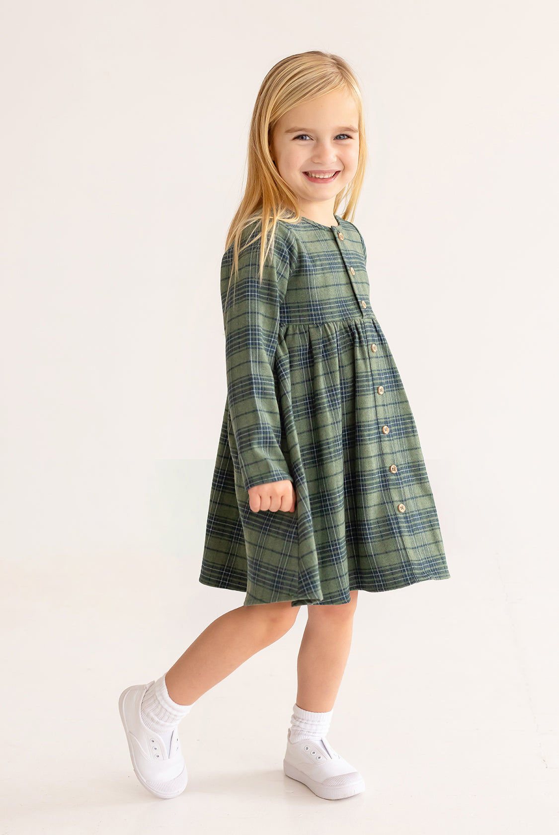 A young girl with long blonde hair smiles in the Cozy Creatures Dress by Mabel and Honey, featuring green plaid and wooden button details. She pairs it with white socks and sneakers, standing against a plain light background.