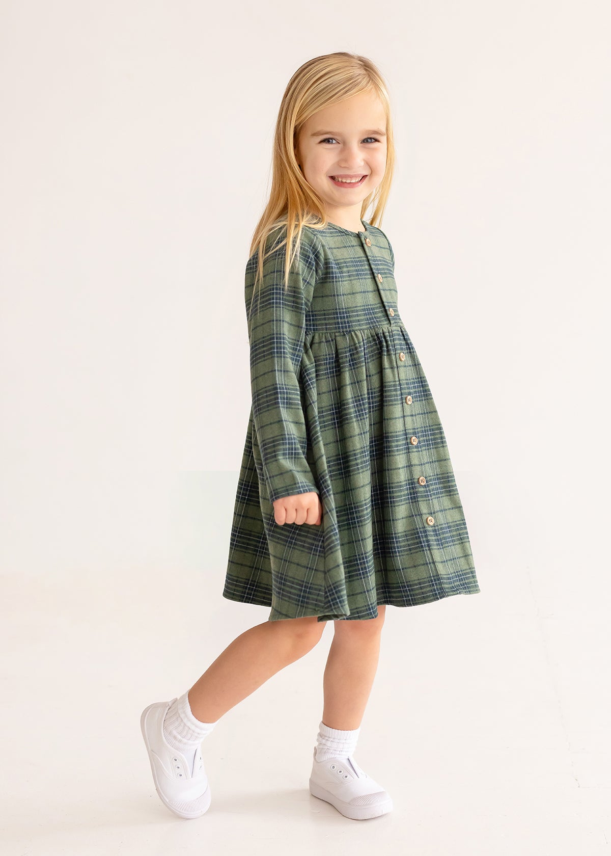 A young girl with long blonde hair smiles in the Cozy Creatures Dress by Mabel and Honey, featuring green plaid and wooden button details. She pairs it with white socks and sneakers, standing against a plain light background.
