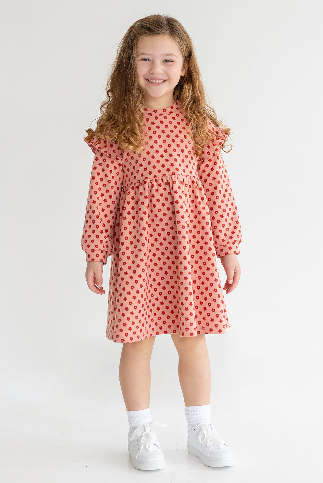 A young girl with long curly hair smiles while wearing the Mabel and Honey Red Velvet Dress, paired with white socks and sneakers, standing against a plain light background.