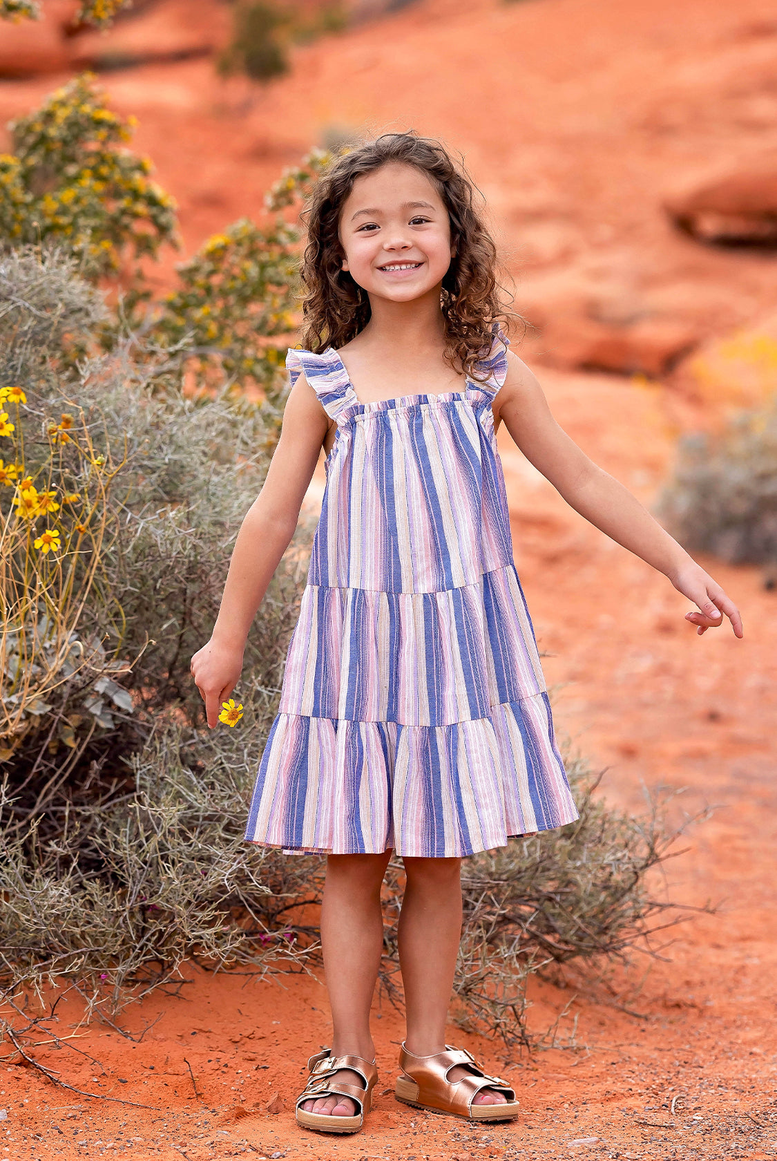 A young girl smiles outdoors on reddish-orange desert soil, surrounded by shrubs and yellow wildflowers, wearing the Riley Rose Dress by Mabel and Honey— a multicolor striped dress with ruffle shoulder straps and sandals.