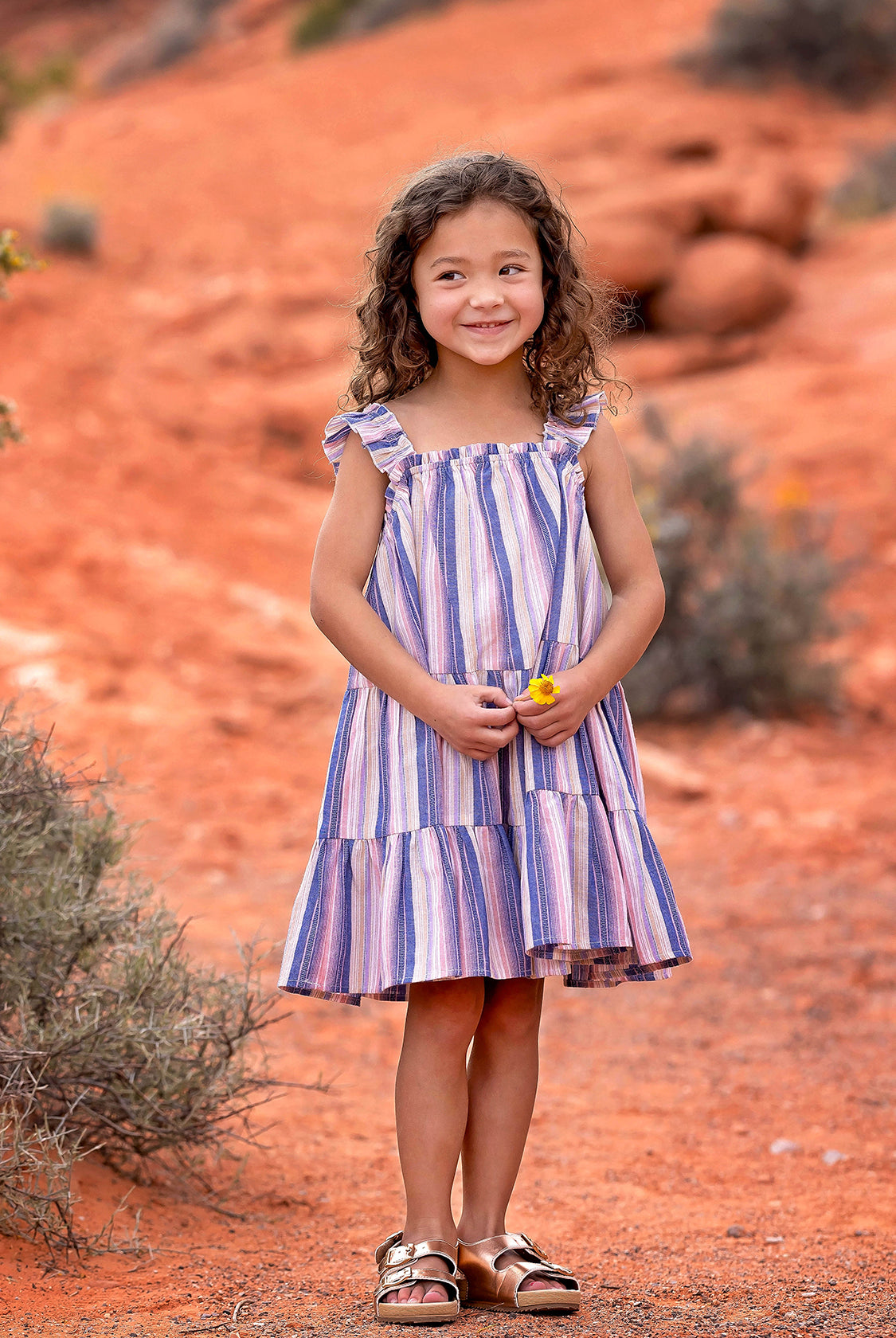 A young girl with curly hair smiles outdoors on red dirt, wearing the Riley Rose Dress by Mabel and Honey. She holds a small yellow flower, with bushes and red rocks in the background.