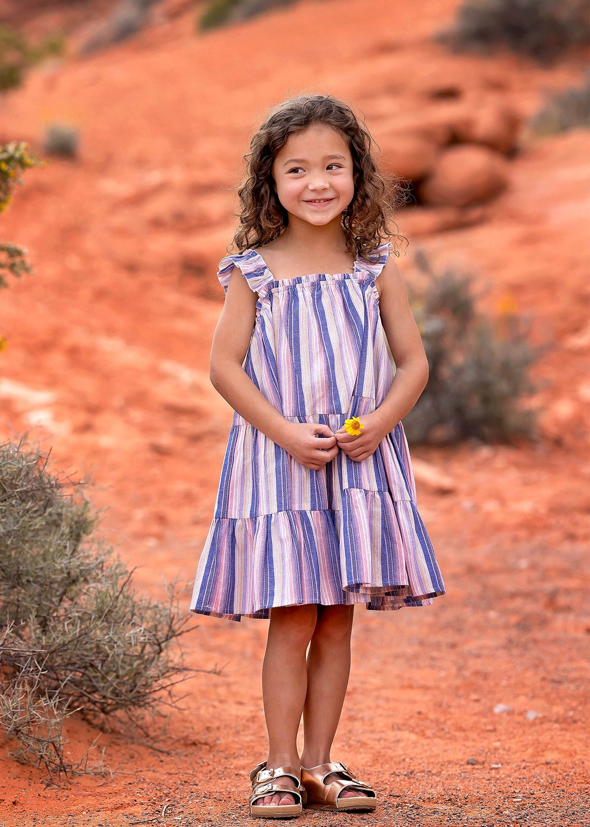 A young girl with curly hair smiles outdoors on red dirt, wearing the Riley Rose Dress by Mabel and Honey. She holds a small yellow flower, with bushes and red rocks in the background.
