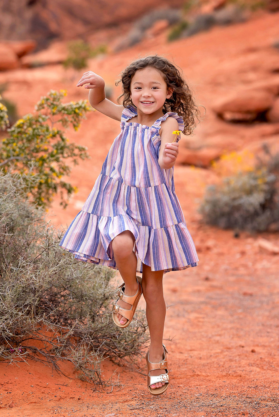 A young girl with curly hair joyfully jumps outdoors in the Riley Rose Dress by Mabel and Honey, featuring multicolor stripes and ruffle shoulder straps, against a red sandy landscape with rocks and shrubs in the background.