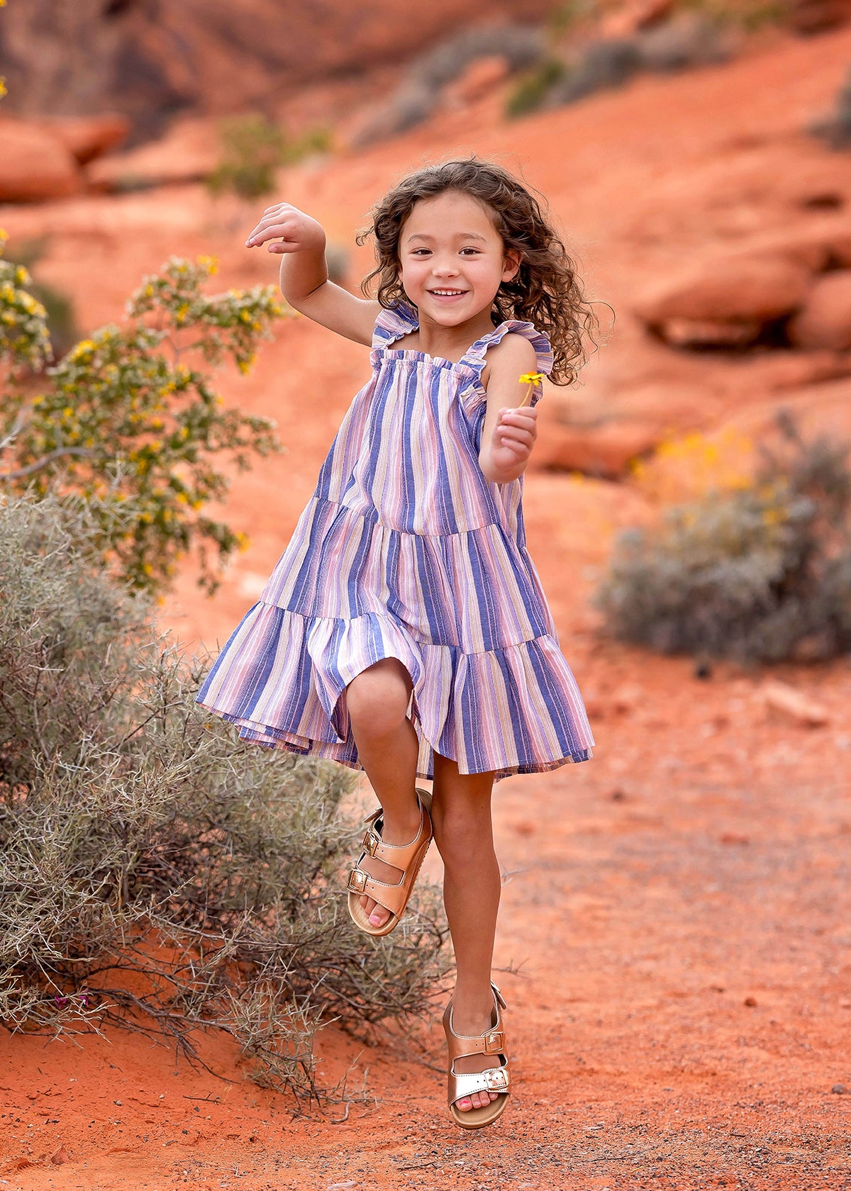 A young girl with curly hair joyfully jumps outdoors in the Riley Rose Dress by Mabel and Honey, featuring multicolor stripes and ruffle shoulder straps, against a red sandy landscape with rocks and shrubs in the background.