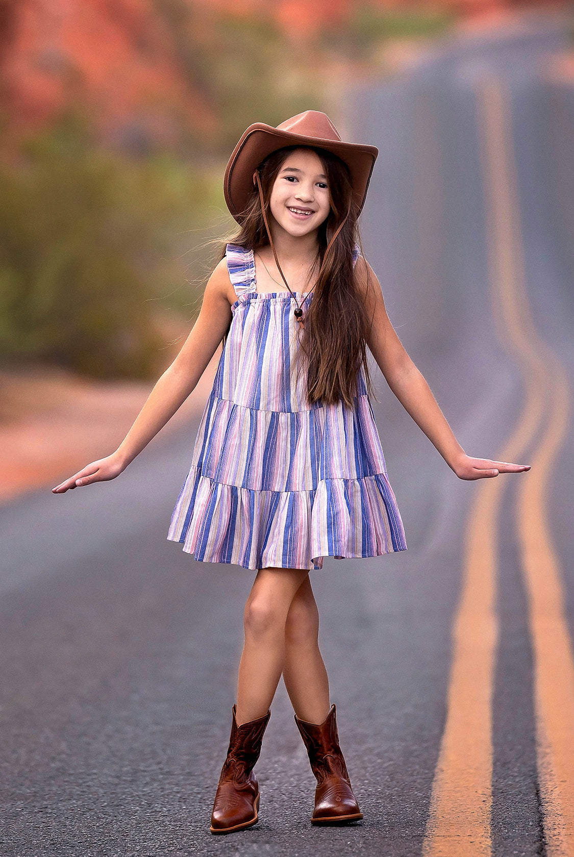 A young girl wears the Mabel and Honey Riley Rose Dress with colorful stripes and ruffle straps, smiling on a winding desert road in brown cowboy boots and a wide-brimmed hat.