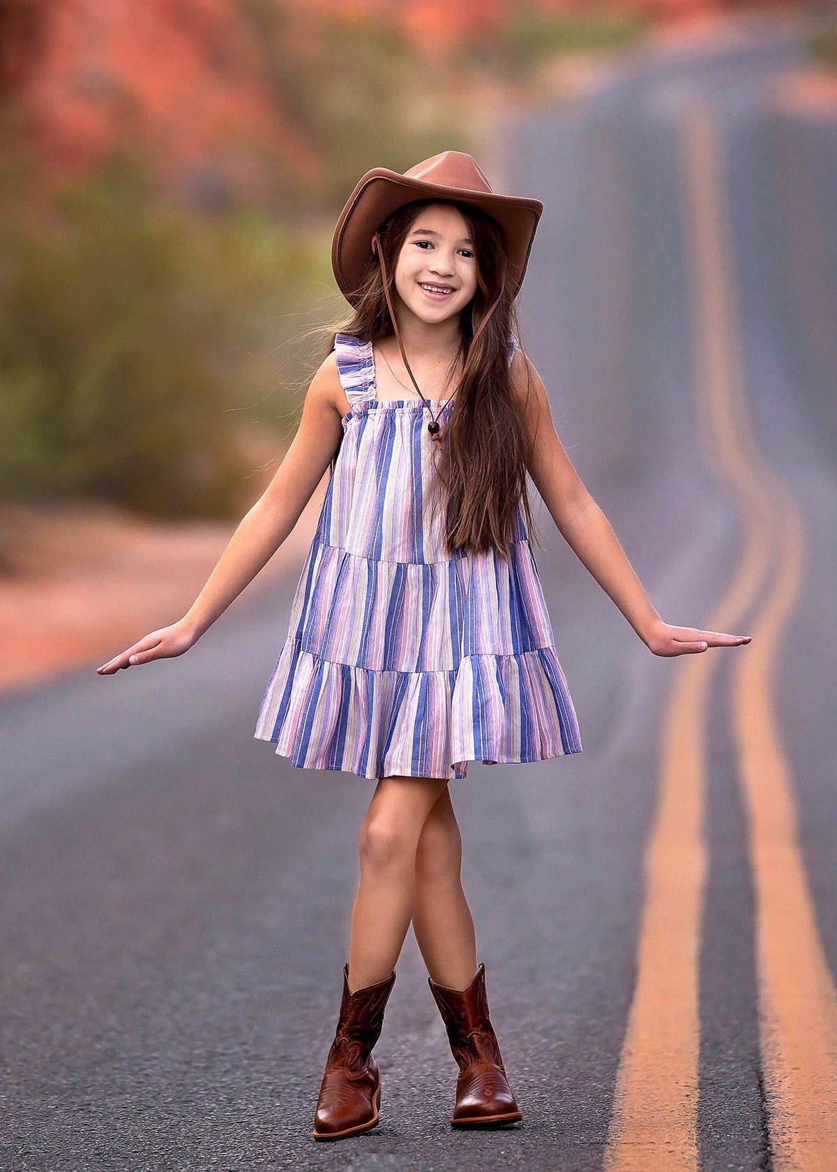 A young girl wears the Mabel and Honey Riley Rose Dress with colorful stripes and ruffle straps, smiling on a winding desert road in brown cowboy boots and a wide-brimmed hat.