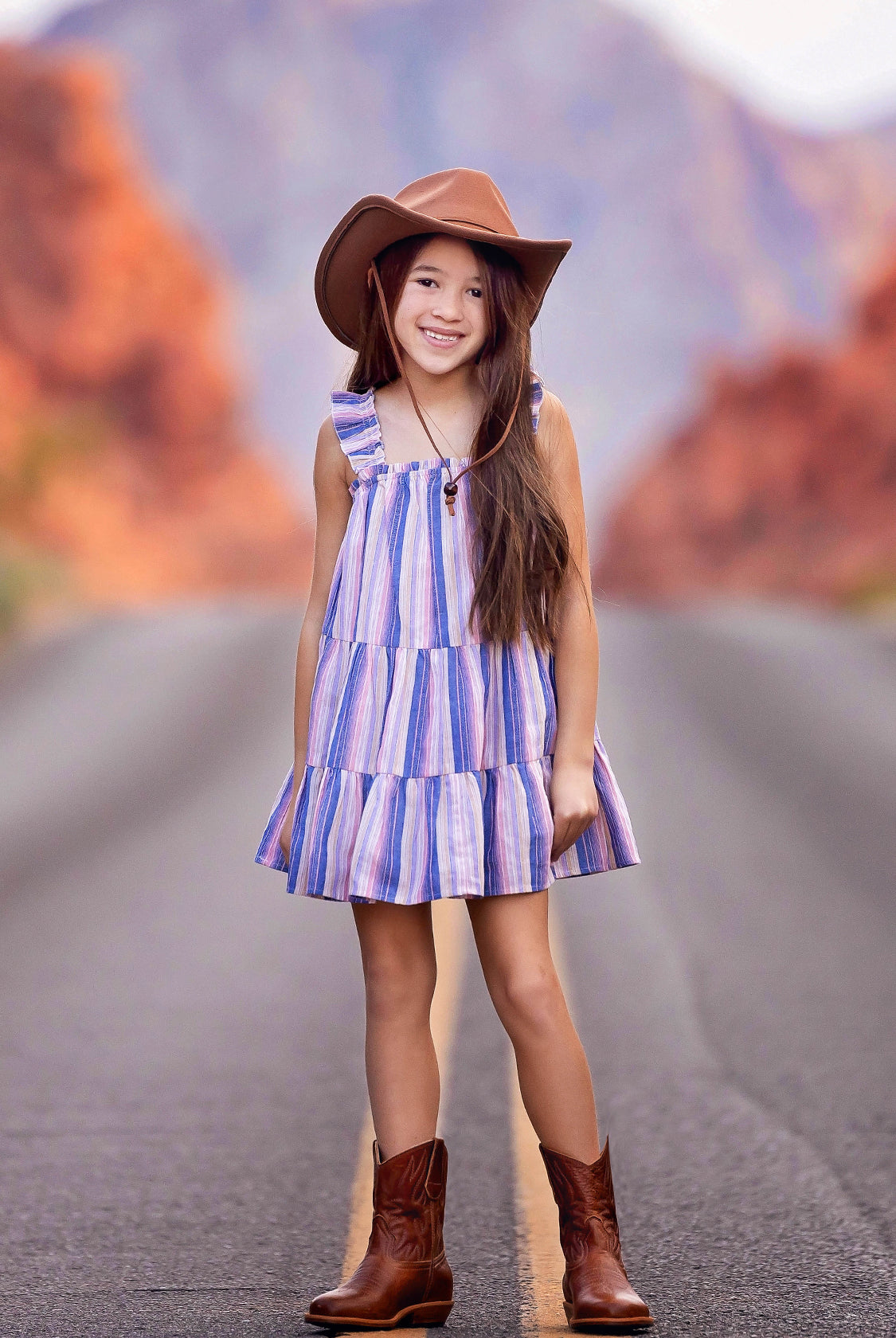 A young girl wearing the Mabel and Honey Riley Rose Dress smiles on a road, paired with a brown cowboy hat and boots. Red rock formations blur in the background.