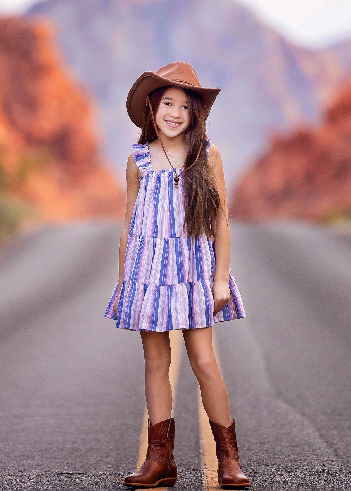 A young girl wearing the Mabel and Honey Riley Rose Dress smiles on a road, paired with a brown cowboy hat and boots. Red rock formations blur in the background.