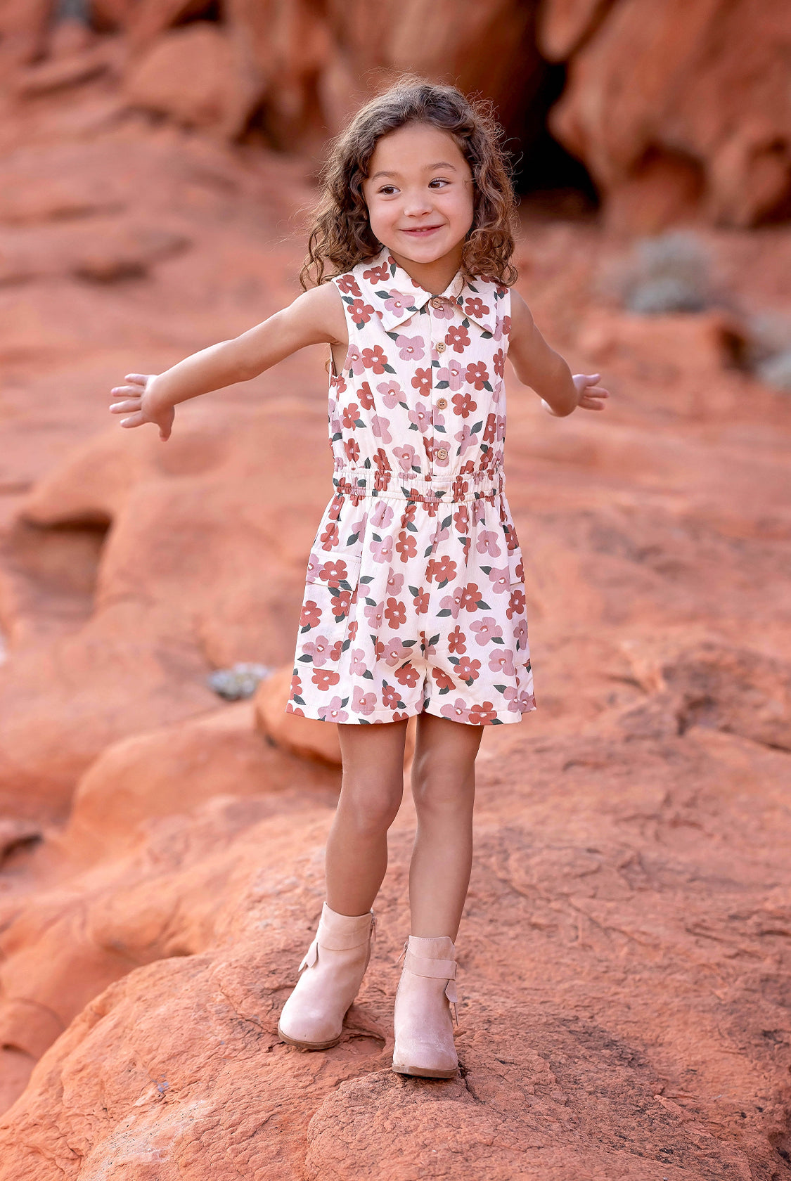 A young child with curly hair smiles and spreads their arms on red rock terrain, wearing the sleeveless collared Mavery Floral Romper by Mabel and Honey with light-colored boots.