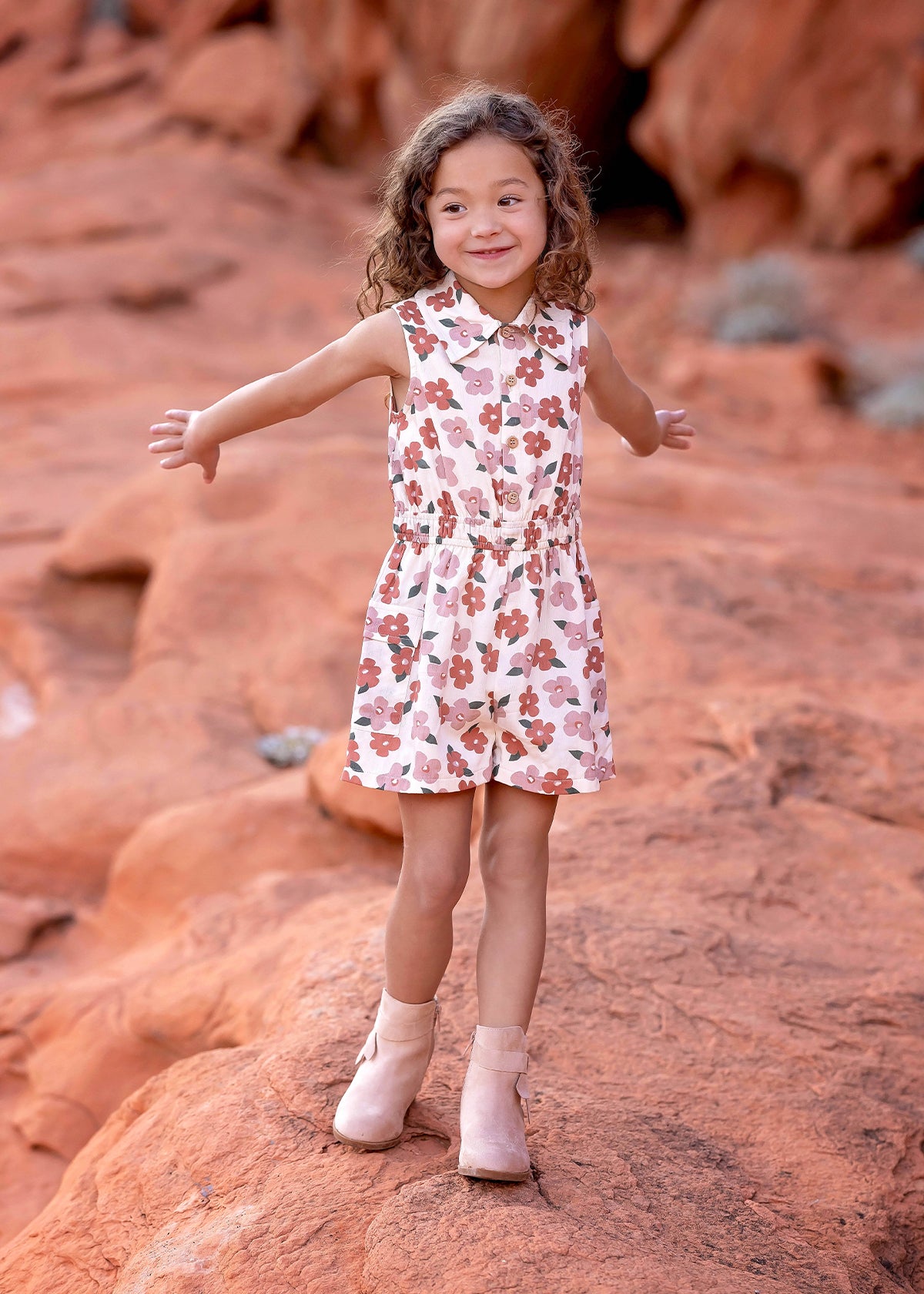 A young girl with curly hair smiles outdoors on red rocks, arms outstretched, wearing the Mavery Floral Romper by Mabel + Honey and light pink boots.