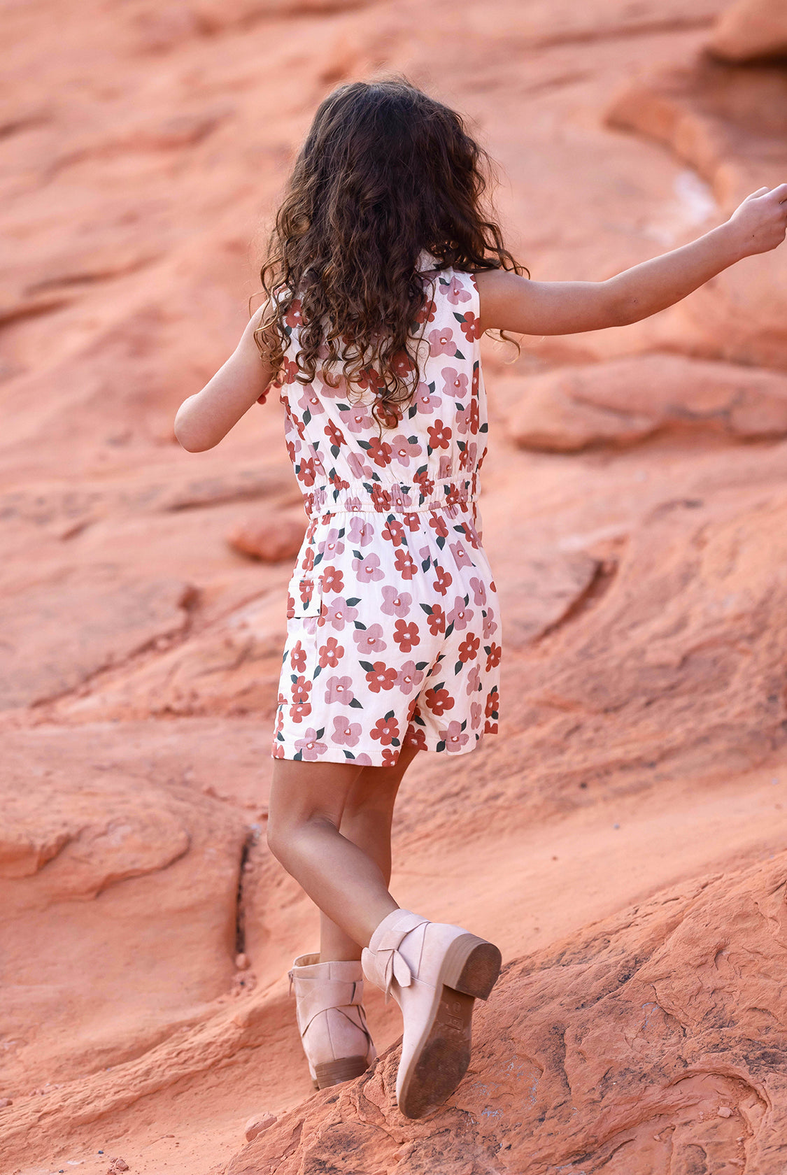 A young girl with curly hair, seen from behind, walks on reddish rocks wearing the Mavery Floral Romper by Mabel and Honey with an elastic waist, paired with light pink boots.